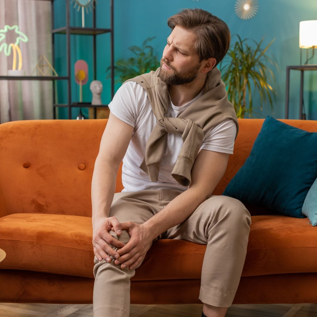 Bearded man with muscle pain holding his knee while sitting on an orange couch