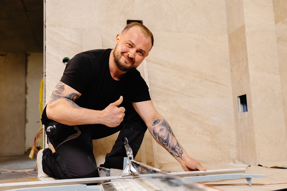 Construction worker cutting beige ceramic tile using manual tile cutter and showing thumbs up, looking satisfied with his work