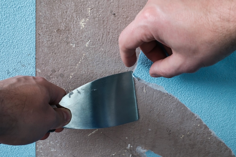 Hand removes old blue wallpaper from the walls, using a spatula. Bathroom renovation. Close-up.